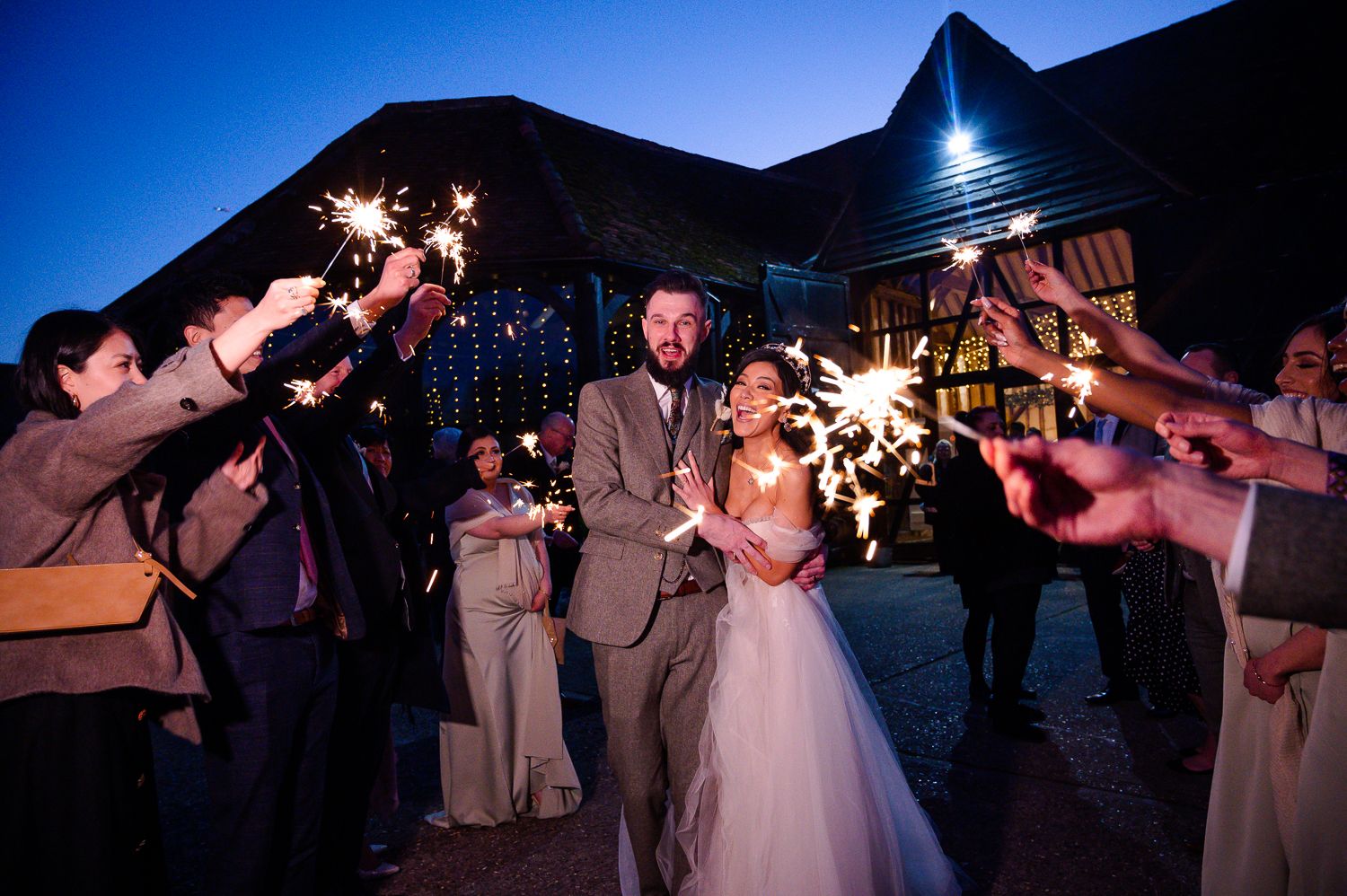Wedding couple celebrates with sparklers surrounded by guests at twilight reception.