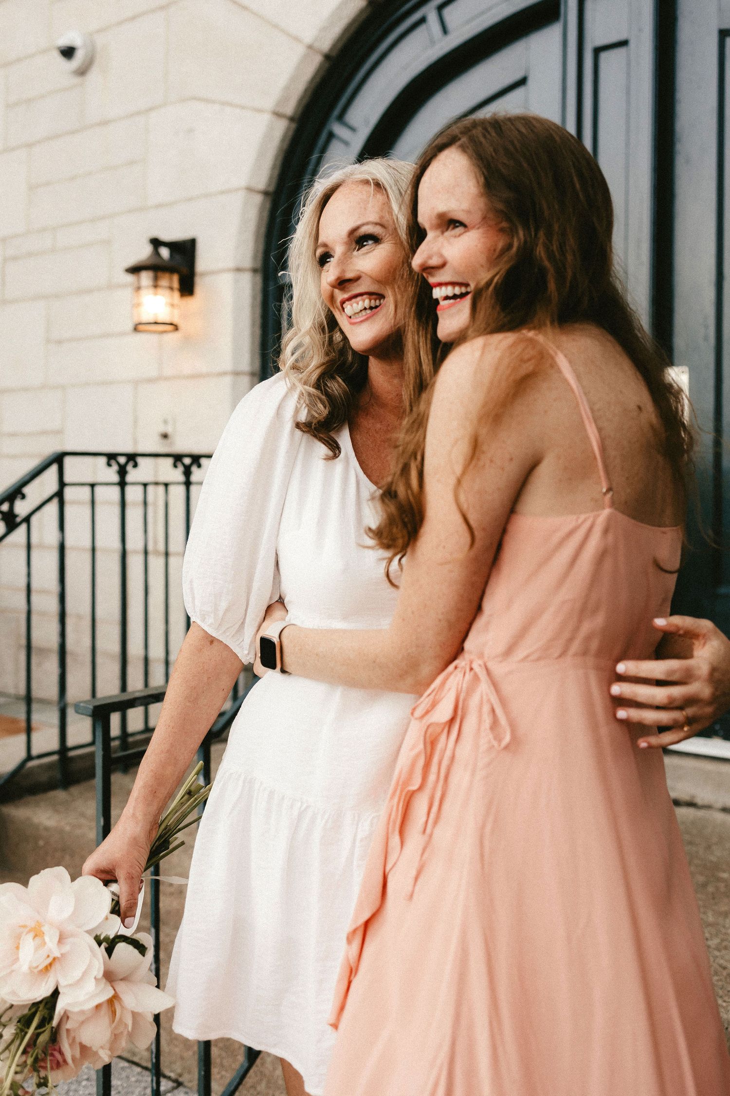 Two women in white and peach dresses share a joyful moment on stone steps.