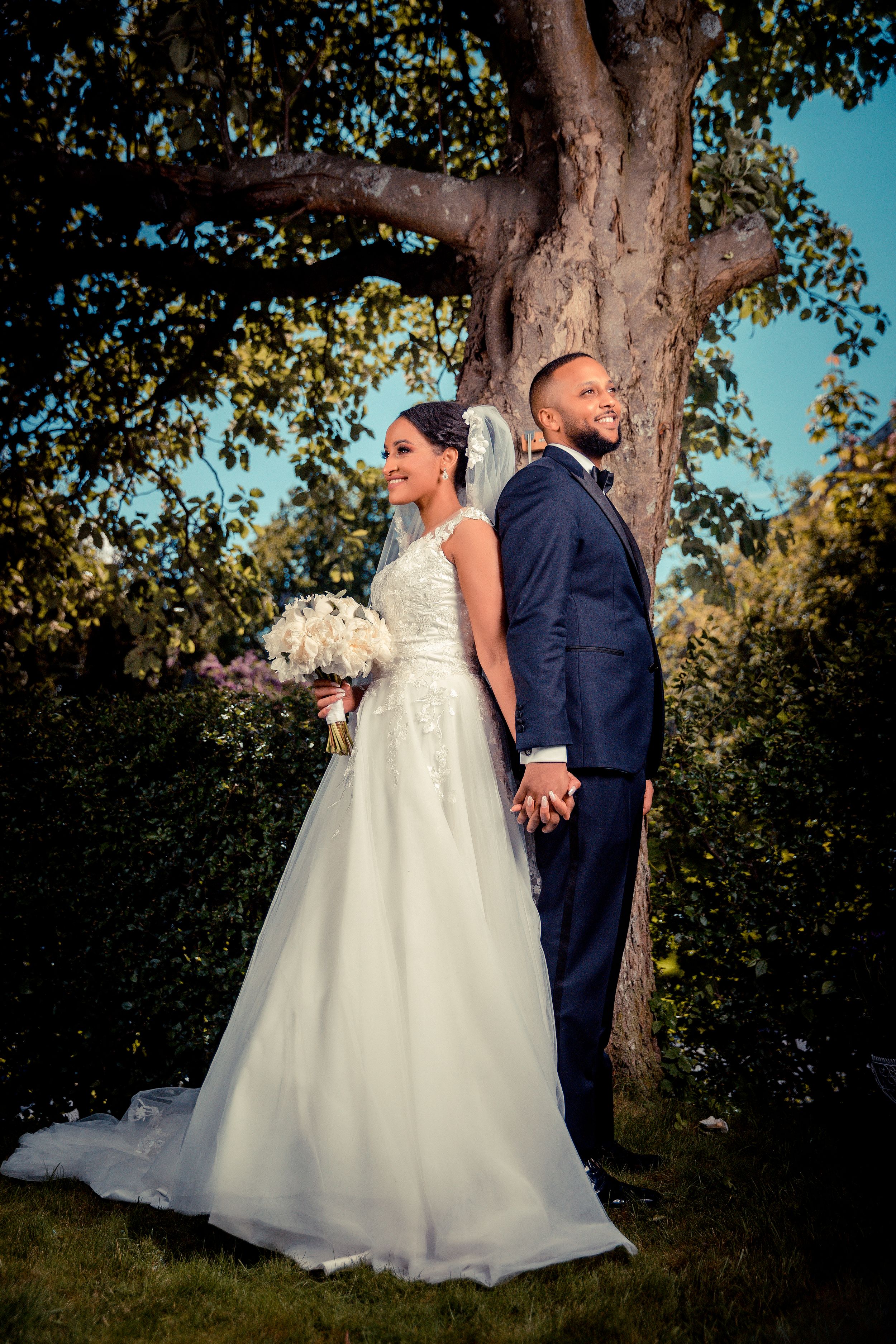 A newlywed couple poses back-to-back under a large tree in formal wedding attire.