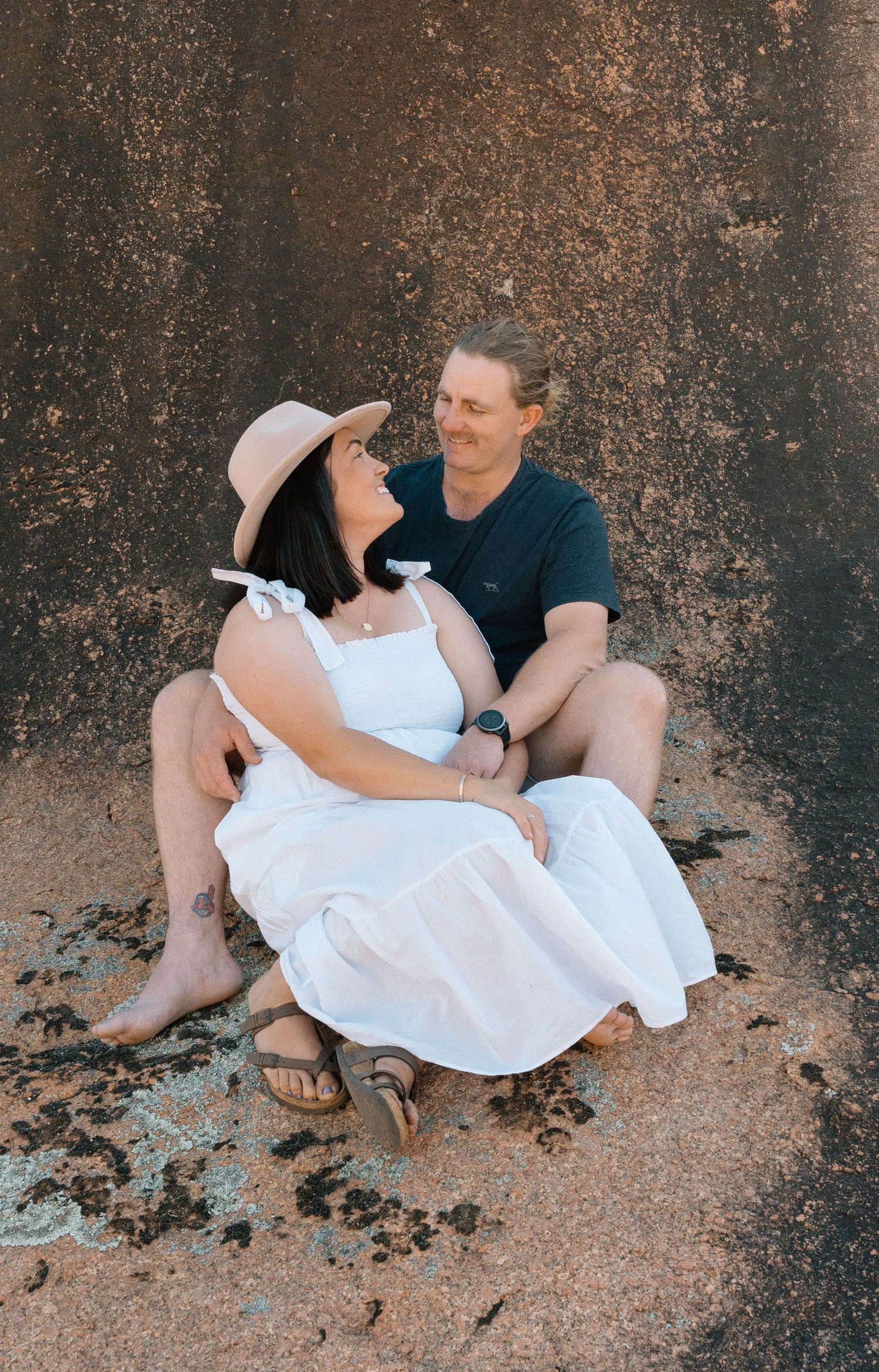 Couple in casual attire sitting against textured wall during outdoor photoshoot.