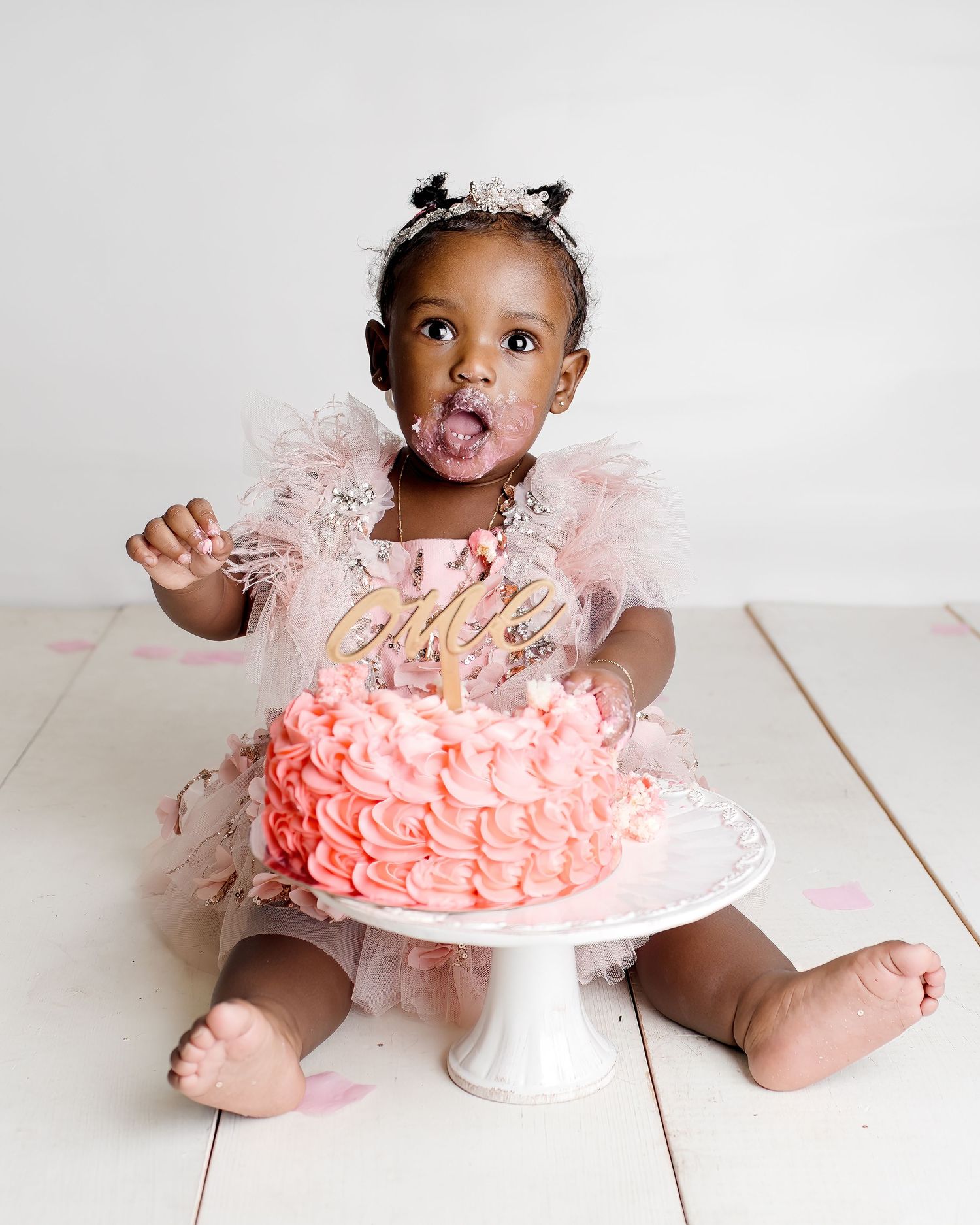 A toddler in a pink tulle dress sits beside a pink frosted birthday cake on a white stand.