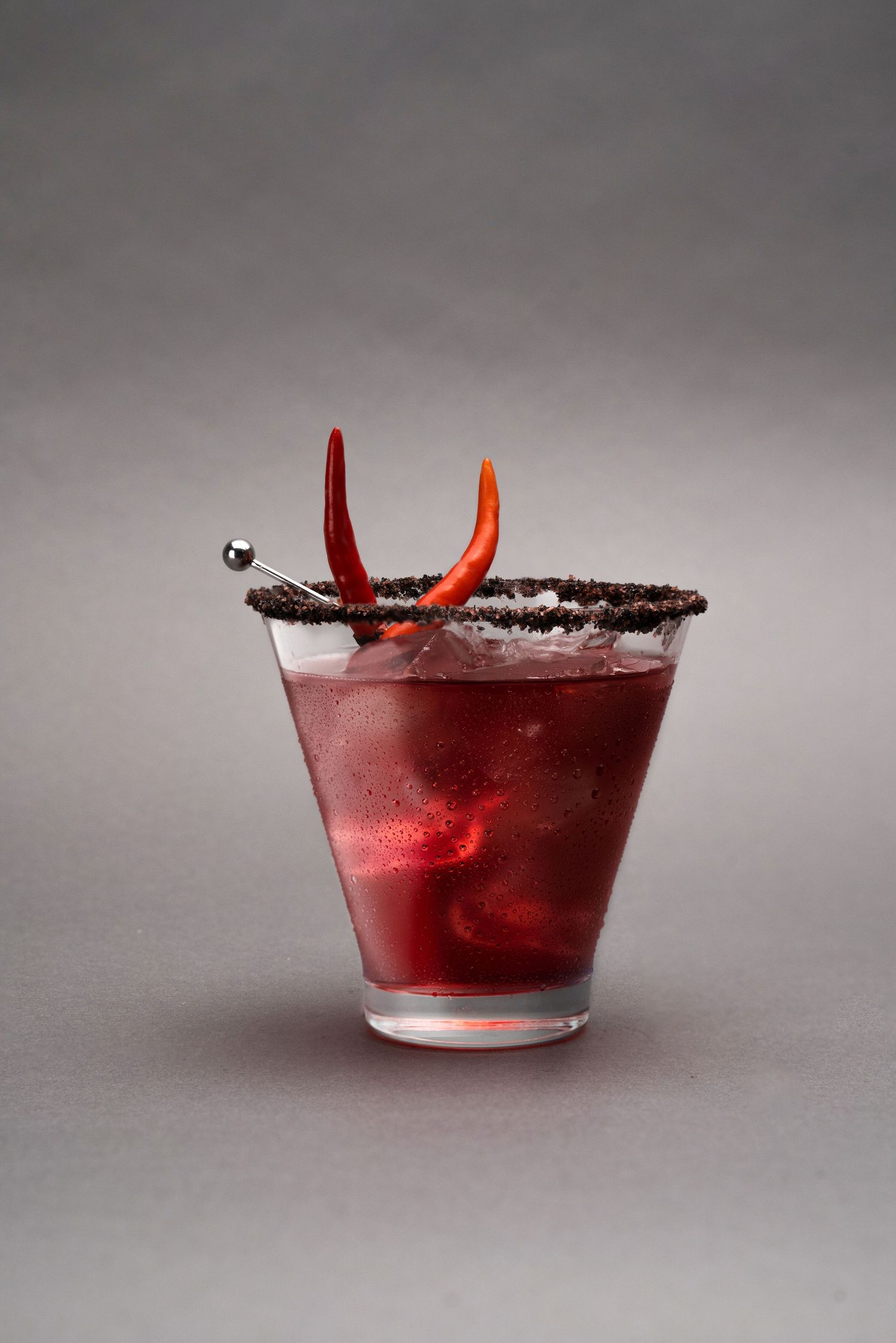 Red cocktail in glass with salt rim and pepper garnish on gray background.