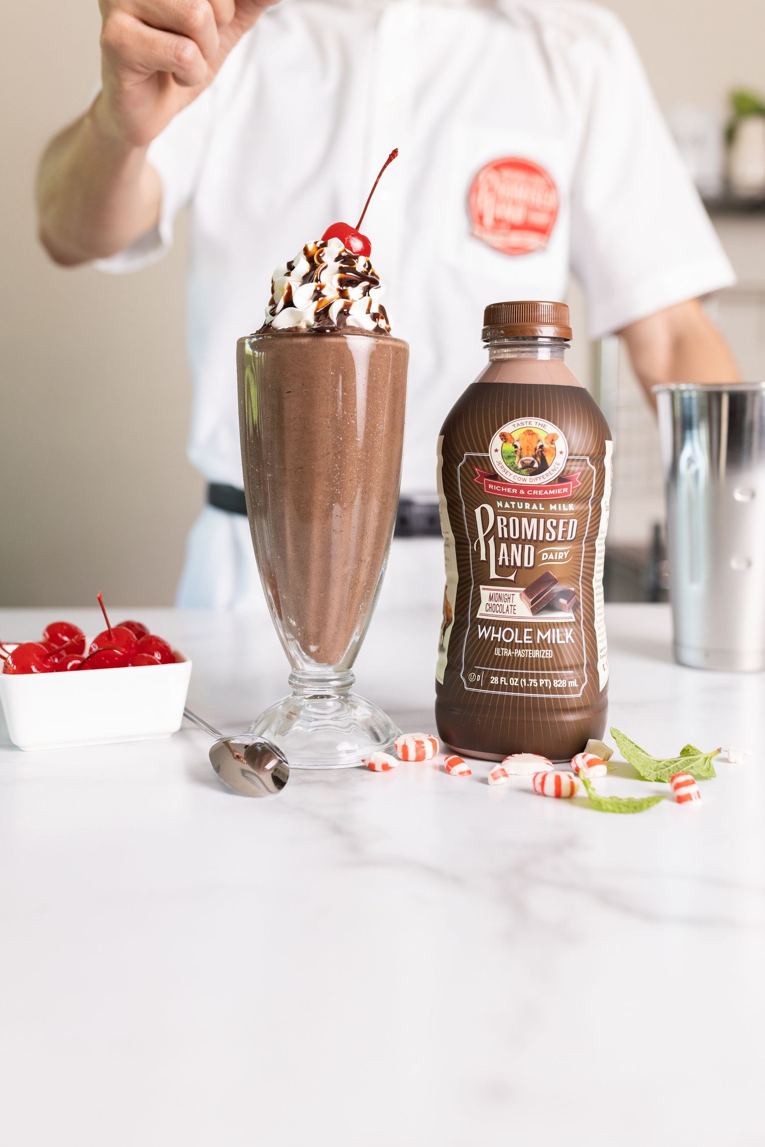 Chocolate milkshakes with whipped cream and cherries alongside Promise Land chocolate milk bottles.
