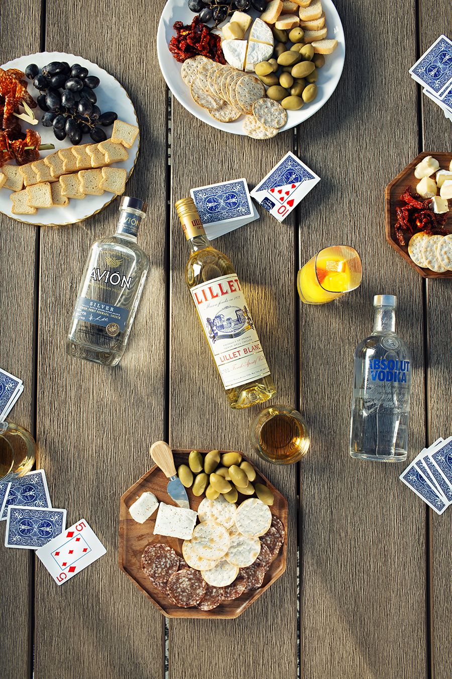 Charcuterie board with snacks, playing cards and drinks spread across wooden deck.