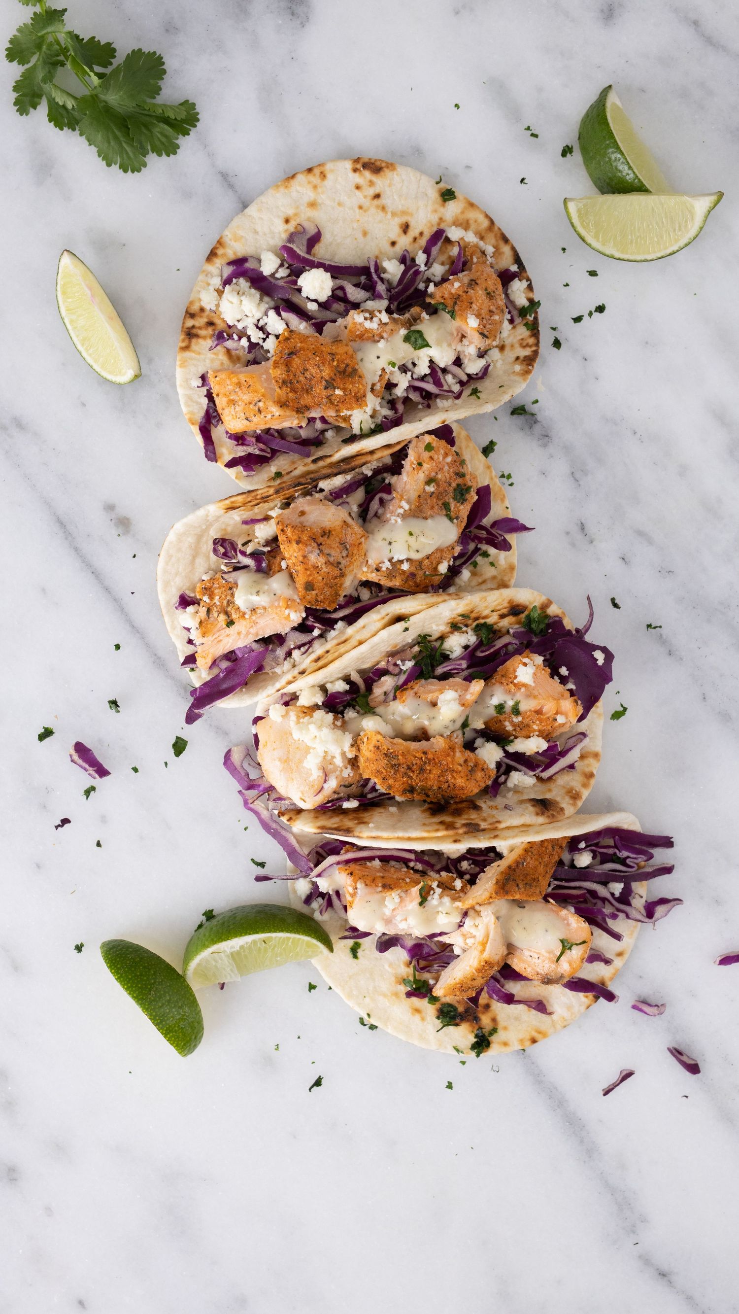 Fish tacos garnished with purple cabbage, cilantro and lime wedges on marble surface.