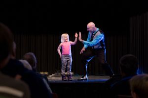Young blonde girl high-fiving a bald magician on a performance stage.