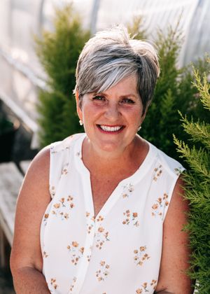 Smiling older woman with short gray hair in floral blouse posing near evergreen trees.