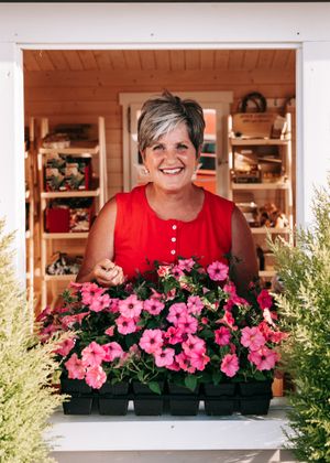 Smiling woman in red top presenting pink petunias at garden shop window display.