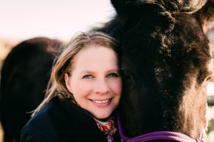 Smiling woman with blonde hair affectionately pressing her cheek against a dark horse outdoors.