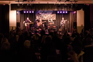 Live band performing on a lit stage to an enthusiastic crowd at an indoor venue.