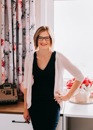 Stylish blonde woman in glasses and white cardigan leaning against desk in bright office.