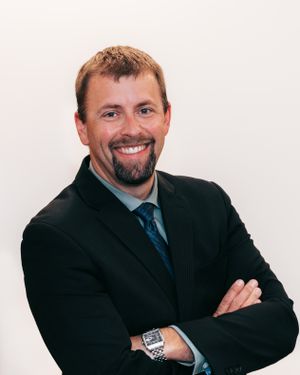 Professional headshot of a smiling man with a beard wearing a dark suit and tie with arms crossed.
