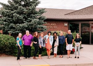 Group of ten people posing outdoors in front of the Luseland Press 331 brick building.