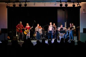 Large group of musicians performing on a lit stage for a seated audience.
