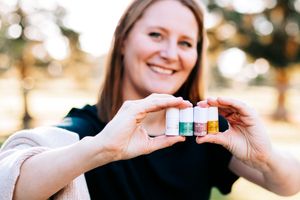 Smiling woman holding four small colorful essential oil roller bottles outdoors in a park.
