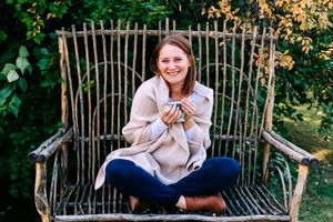 Smiling woman sitting cross-legged on a rustic wooden bench holding a mug outdoors.