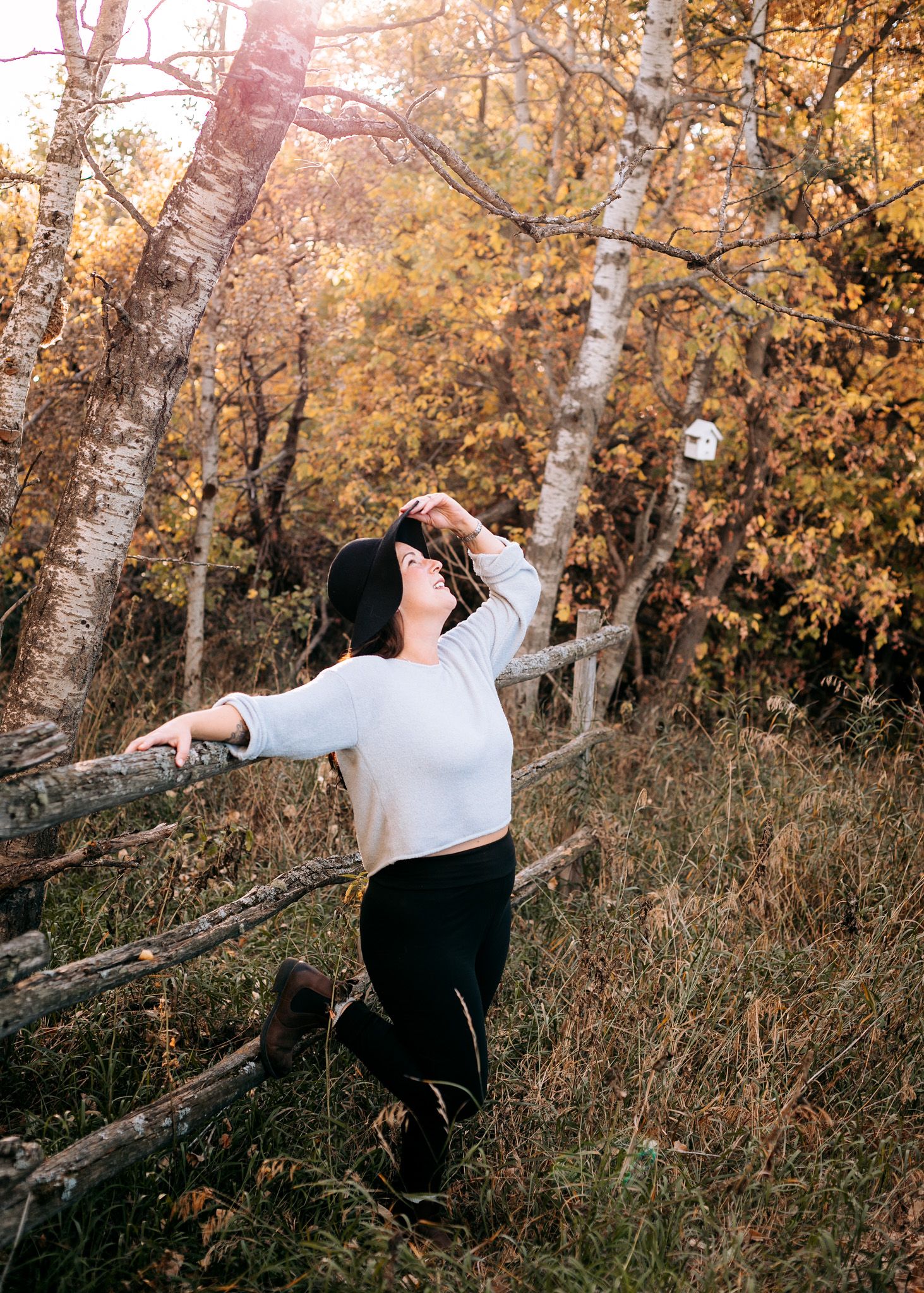Woman in white top leaning on rustic fence in golden autumn forest.