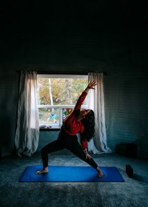 Woman practicing warrior yoga pose on blue mat in dimly lit room by window.
