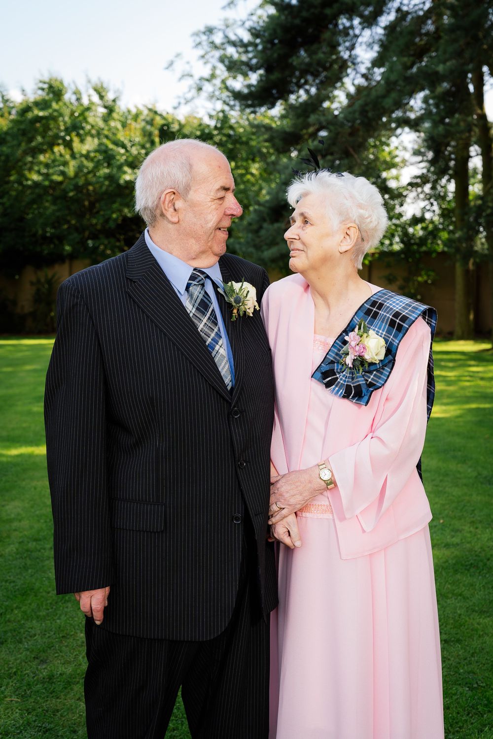 Elderly couple in formal attire sharing tender moments outdoors, with the woman wearing a pink dress.