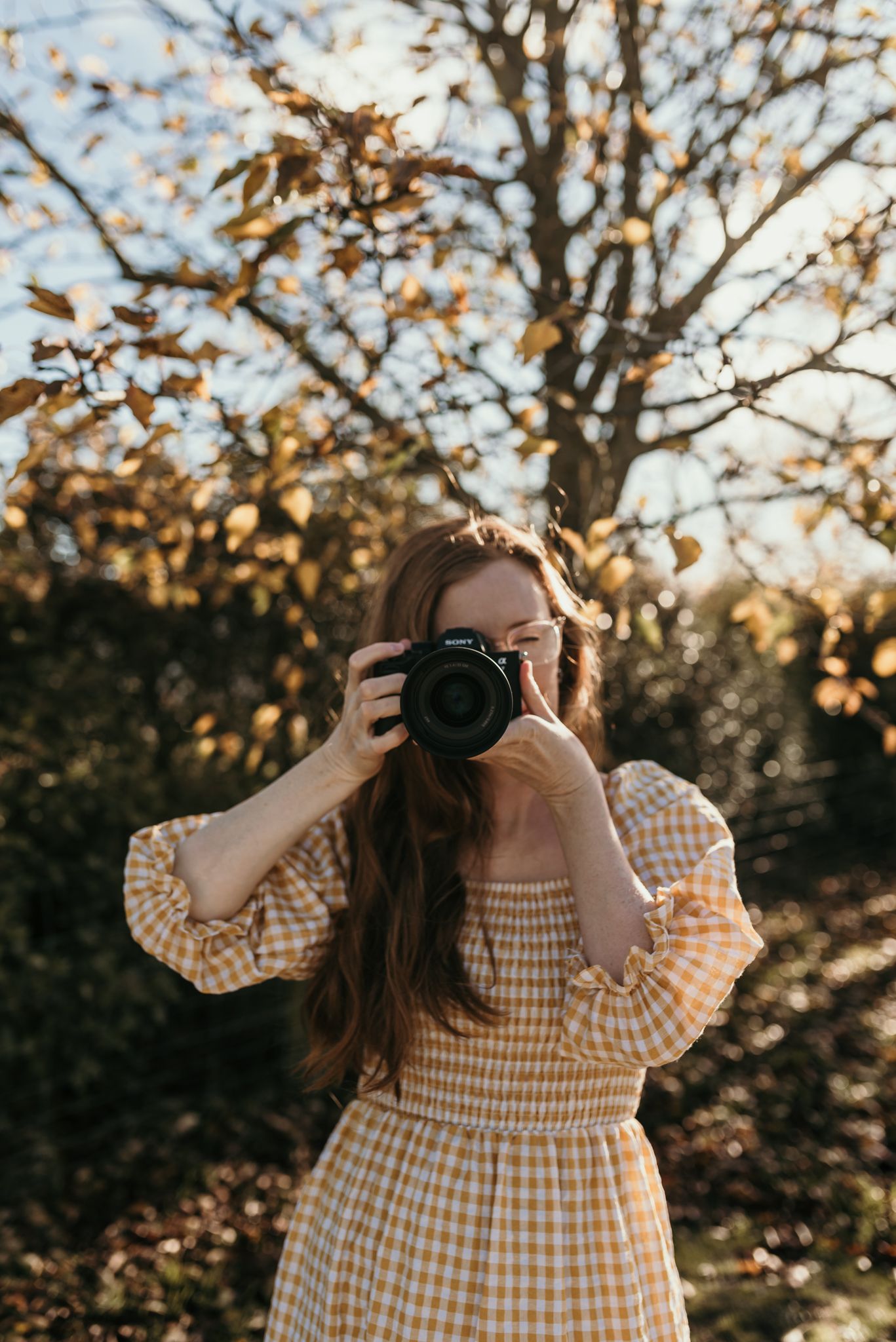 Photographer in yellow gingham dress taking photos in autumn woodland setting during golden hour.