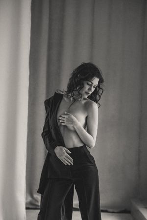Monochrome portrait in loose-fitting pants and open jacket against curtained backdrop.