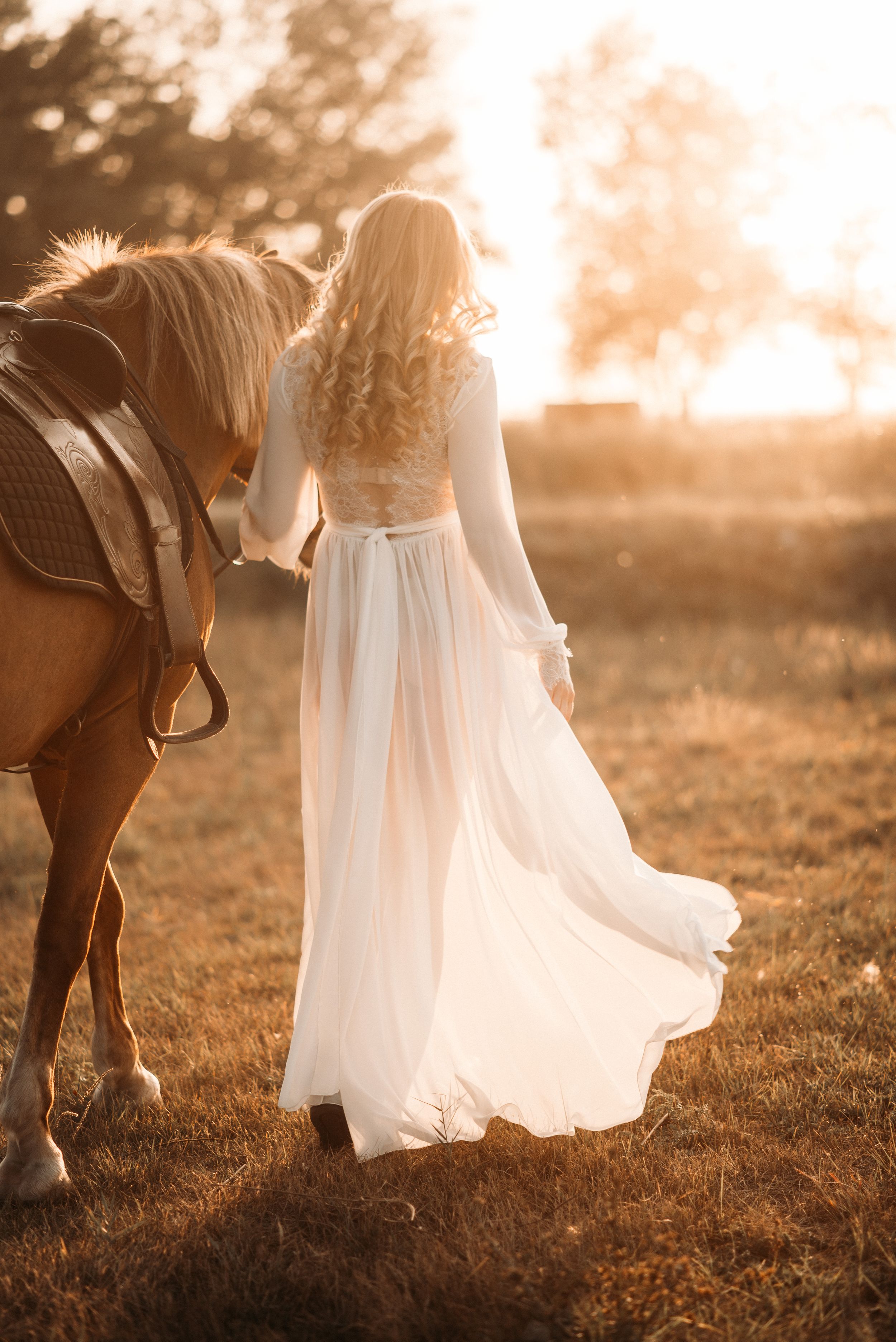 Flowing white dress photographed in golden sunlight with a horse in a dreamy outdoor setting.
