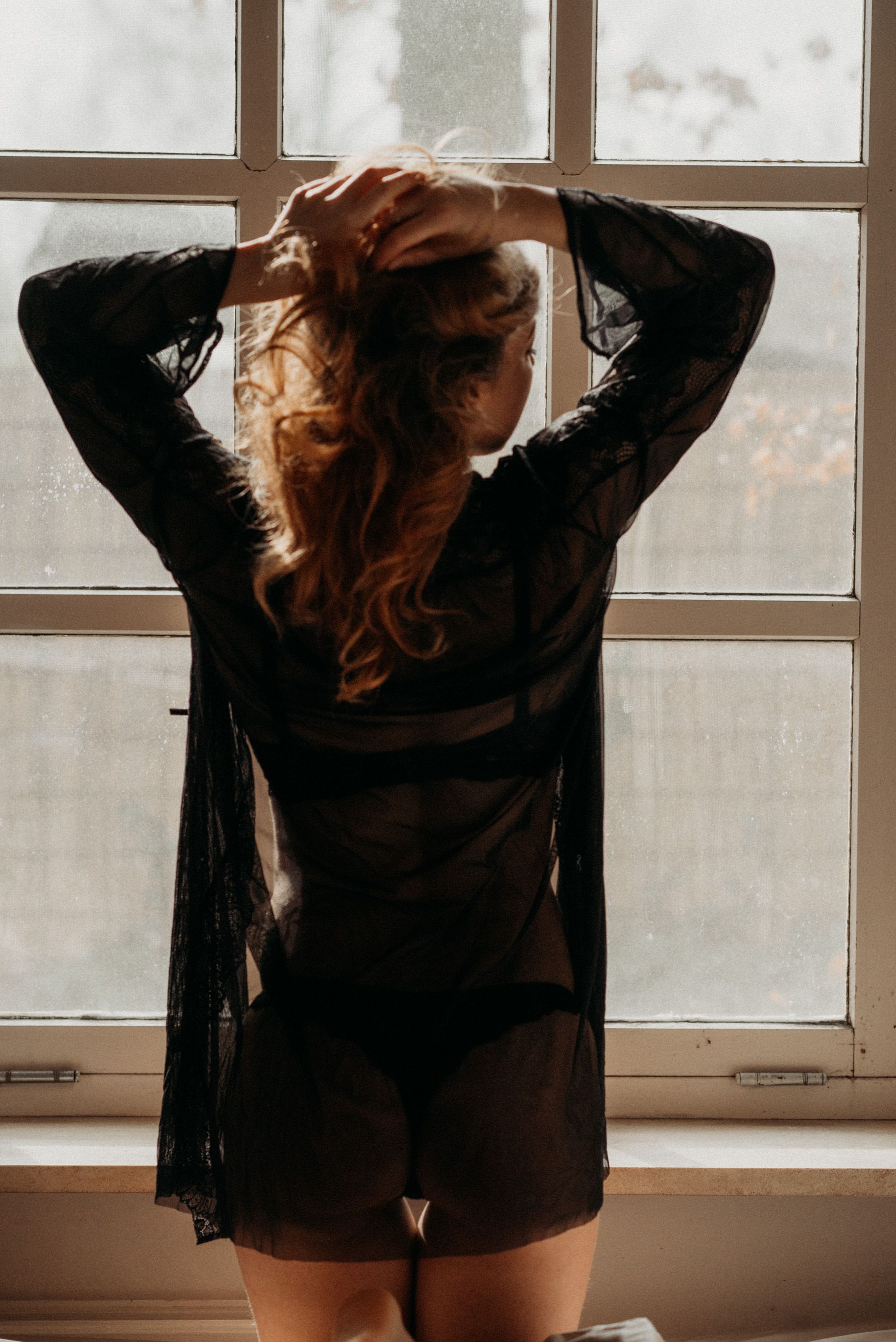 Person in sheer clothing poses by window in natural morning light.