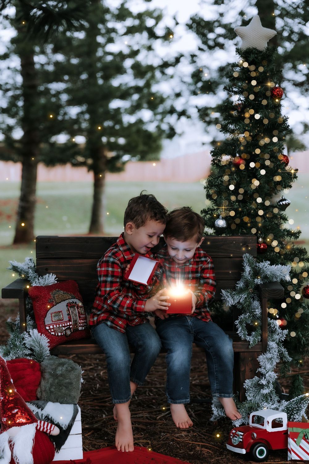 Two children in plaid shirts share a magical moment with glowing light on a bench by a Christmas tree.