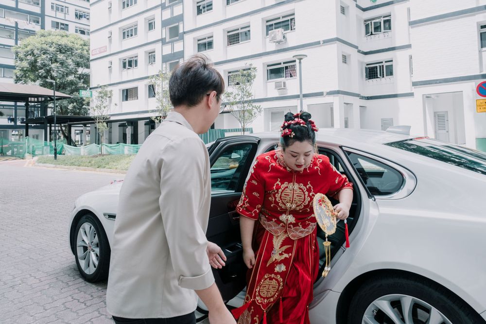 A person in a gray suit helps someone in traditional red Chinese dress exit a car.