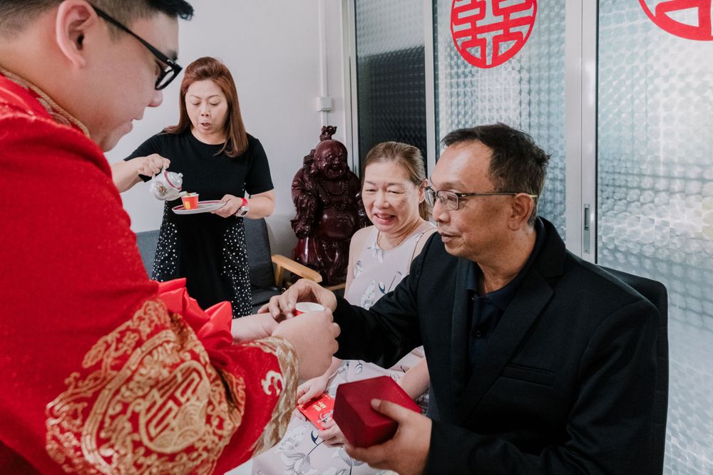 A tea ceremony participant in red traditional dress serves tea to seated family members.