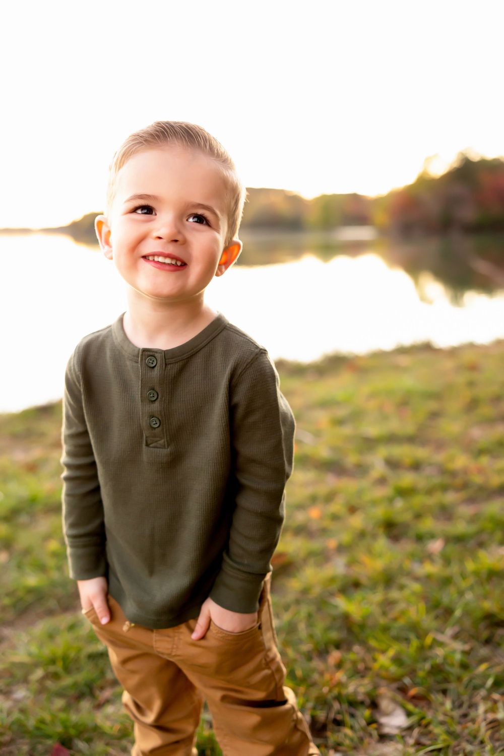 Child wearing an olive henley shirt smiles outdoors at sunset by a lake.