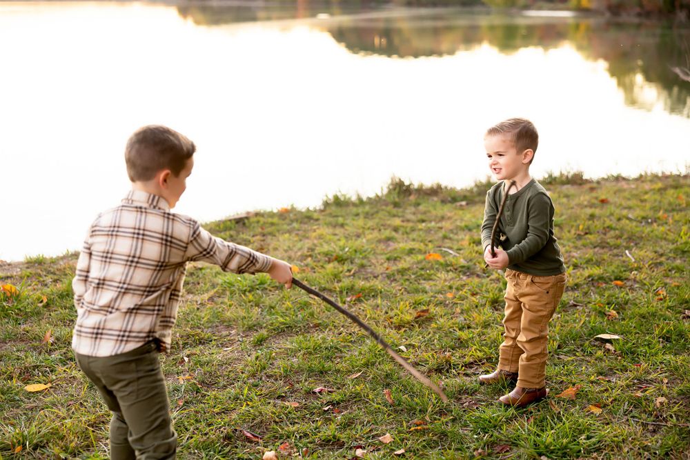 Two children playing with sticks near a lake during golden hour in a grassy area.