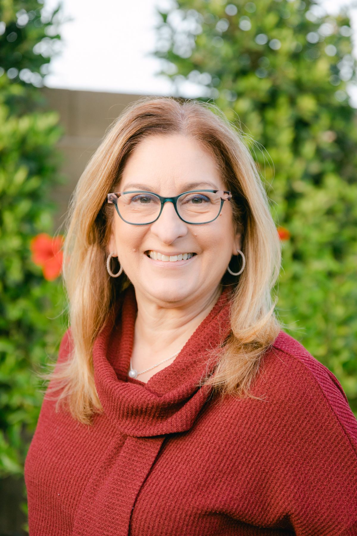A woman with blonde hair and a red sweater smiles at the camera. She has glasses and there are hibiscus flowers and bushes behind her.