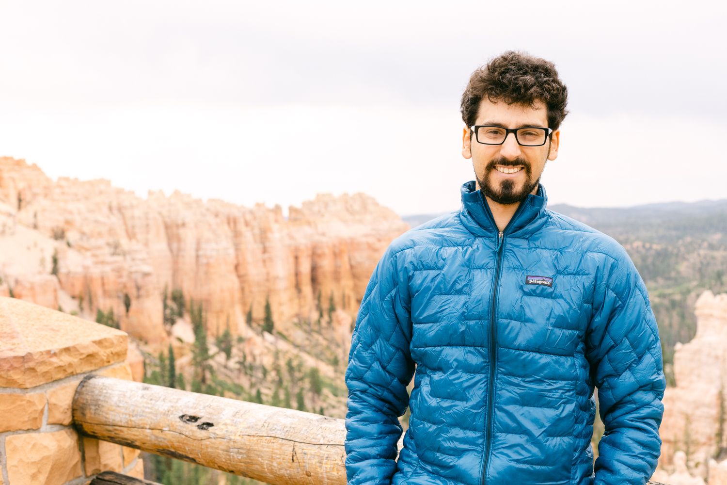 A man with brown curly hair stands at an overlook in Bryce Canyon National Park. He's in a blue jacket. There are orange hoodoos behind him.