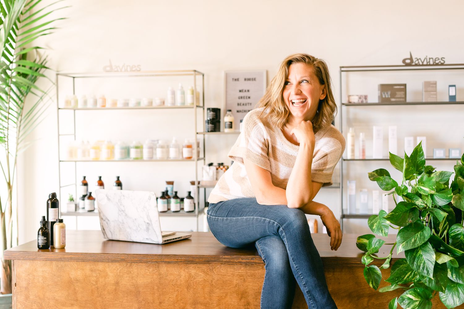 A blonde woman sits on a counter and laughs. She's in a striped shirt and there are hair products on shelves in the background.