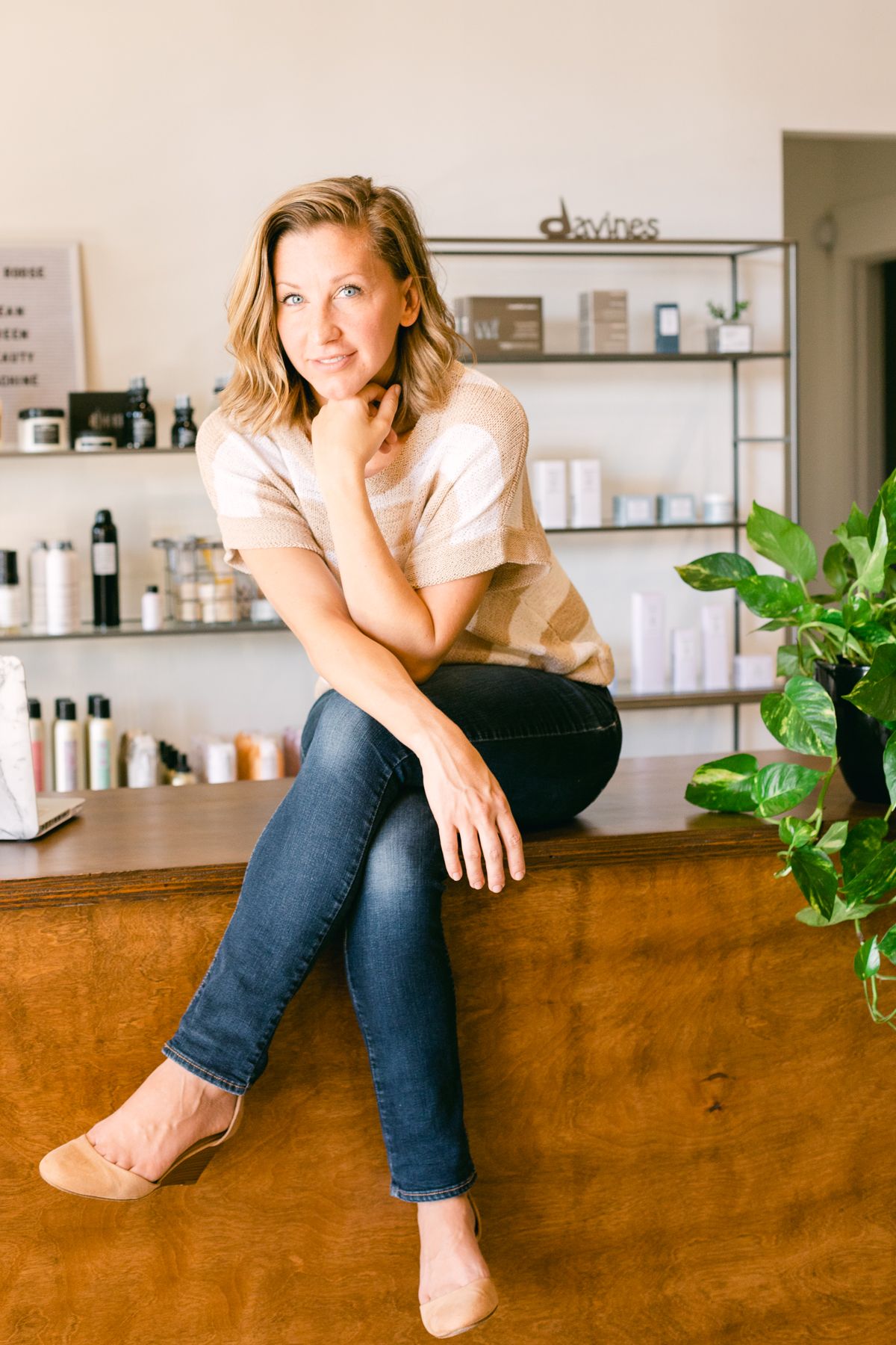 A blonde woman sits on a counter. She's in a striped shirt and there are hair products on shelves in the background.