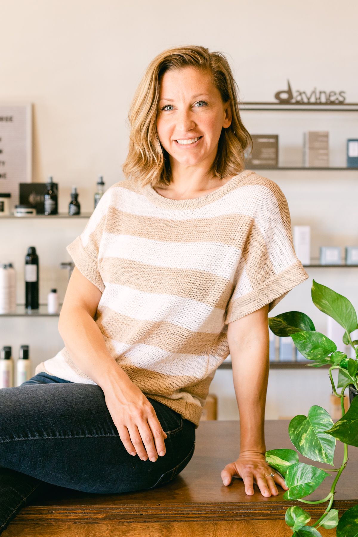 A blonde woman sits on a counter. She's in a striped shirt and there are hair products on shelves in the background.