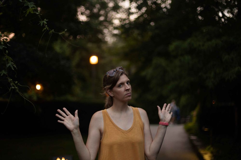 Person in yellow tank top raises hands playfully at dusk with glowing lights in background.