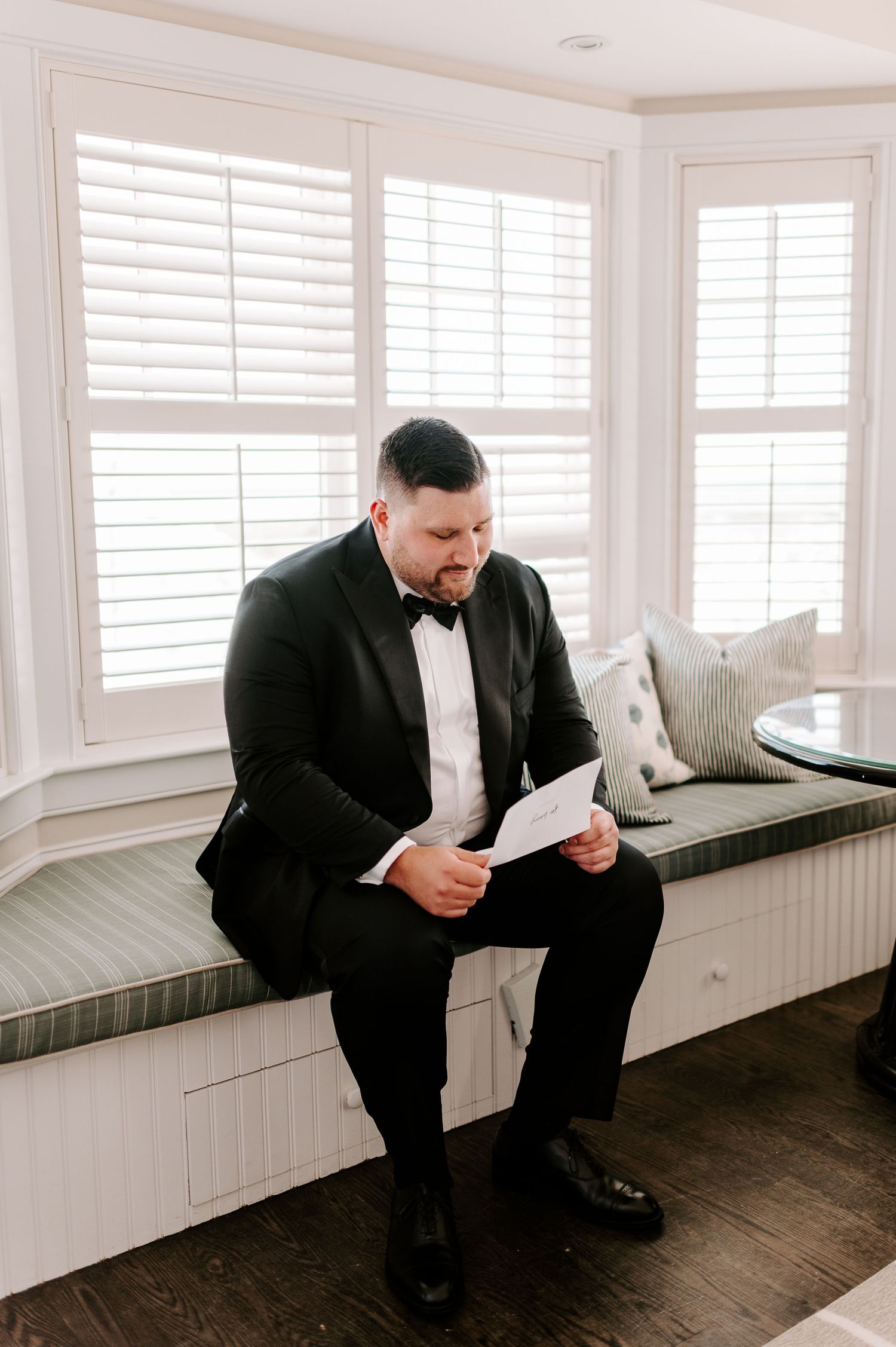 A person in a black tuxedo reads a letter while sitting on a window bench.