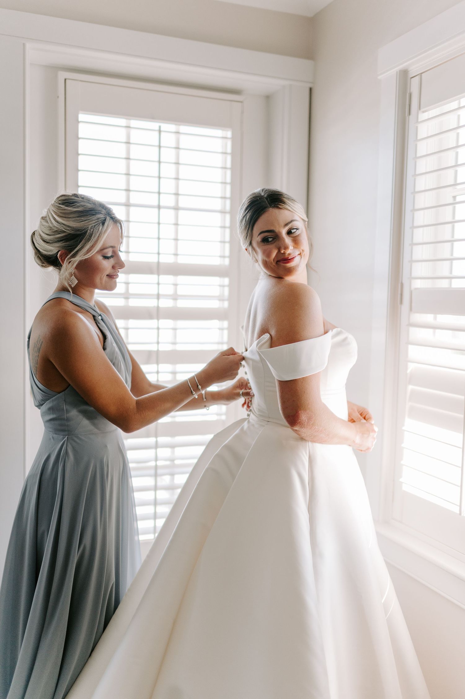 A bridesmaid in blue helps adjust a stunning white wedding gown in natural window light.