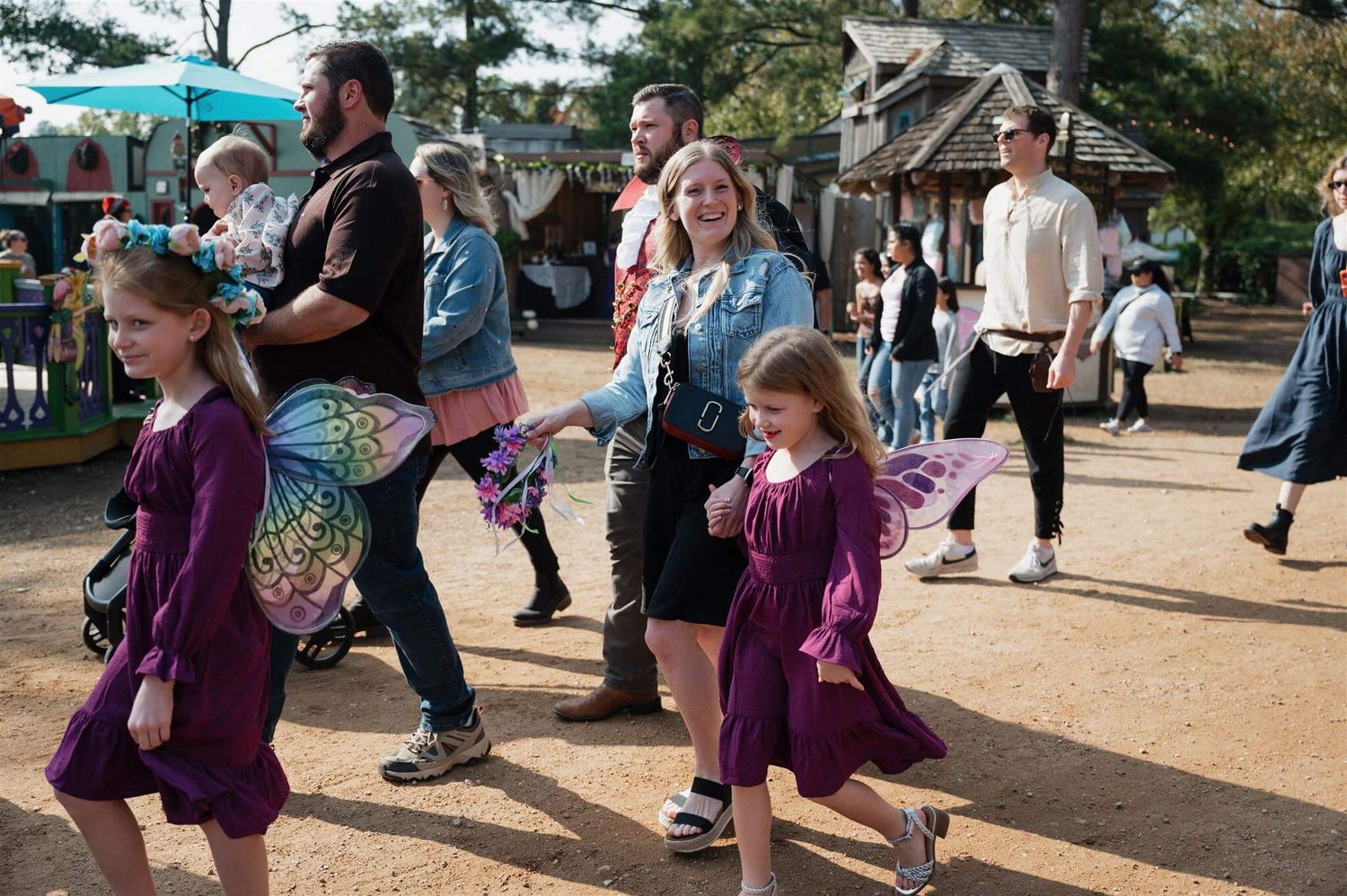 Festival attendees in medieval attire walk along a dirt path at a themed outdoor event.
