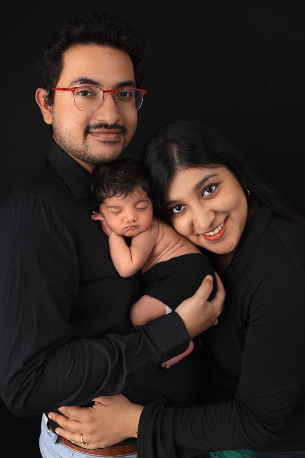 A loving family portrait with parents holding their sleeping newborn baby against a dark backdrop.