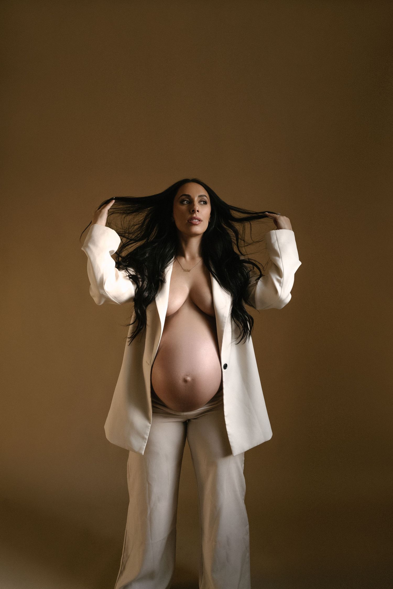 Dynamic maternity portrait with flowing hair and white blazer against brown backdrop.