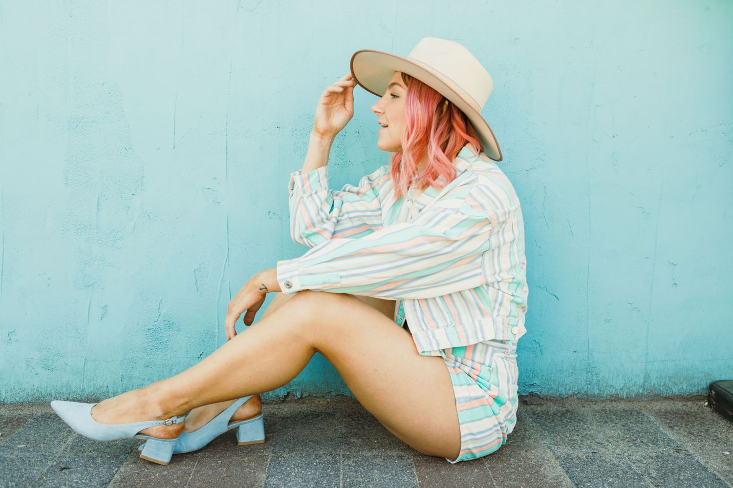 Fashion model in a white hat and summer outfit posing against a turquoise wall.
