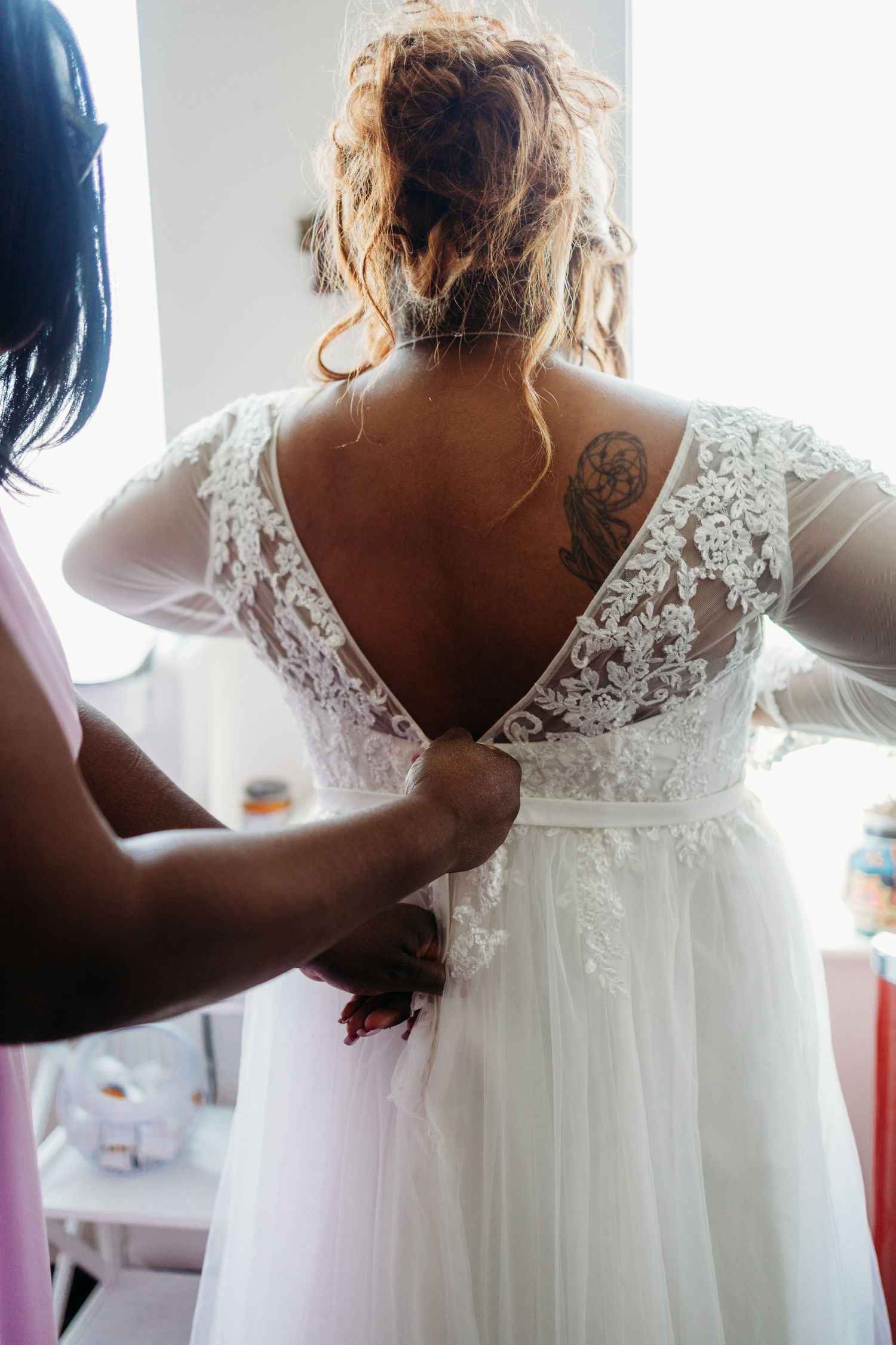 Back view of a white lace wedding dress being buttoned up, showing intricate detail and a visible tattoo.