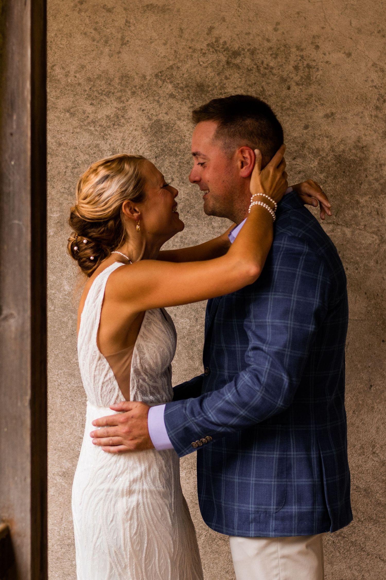 A romantic moment between a couple in wedding attire, with the bride in white and groom in navy blue embracing against a wall.