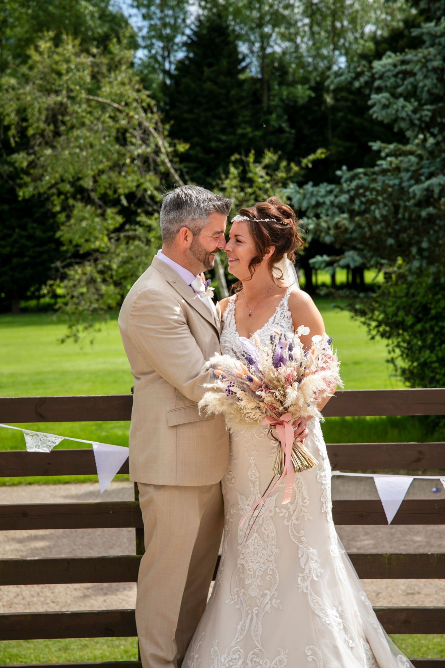 A romantic outdoor wedding scene with a couple sharing a tender moment on a wooden deck surrounded by lush greenery.