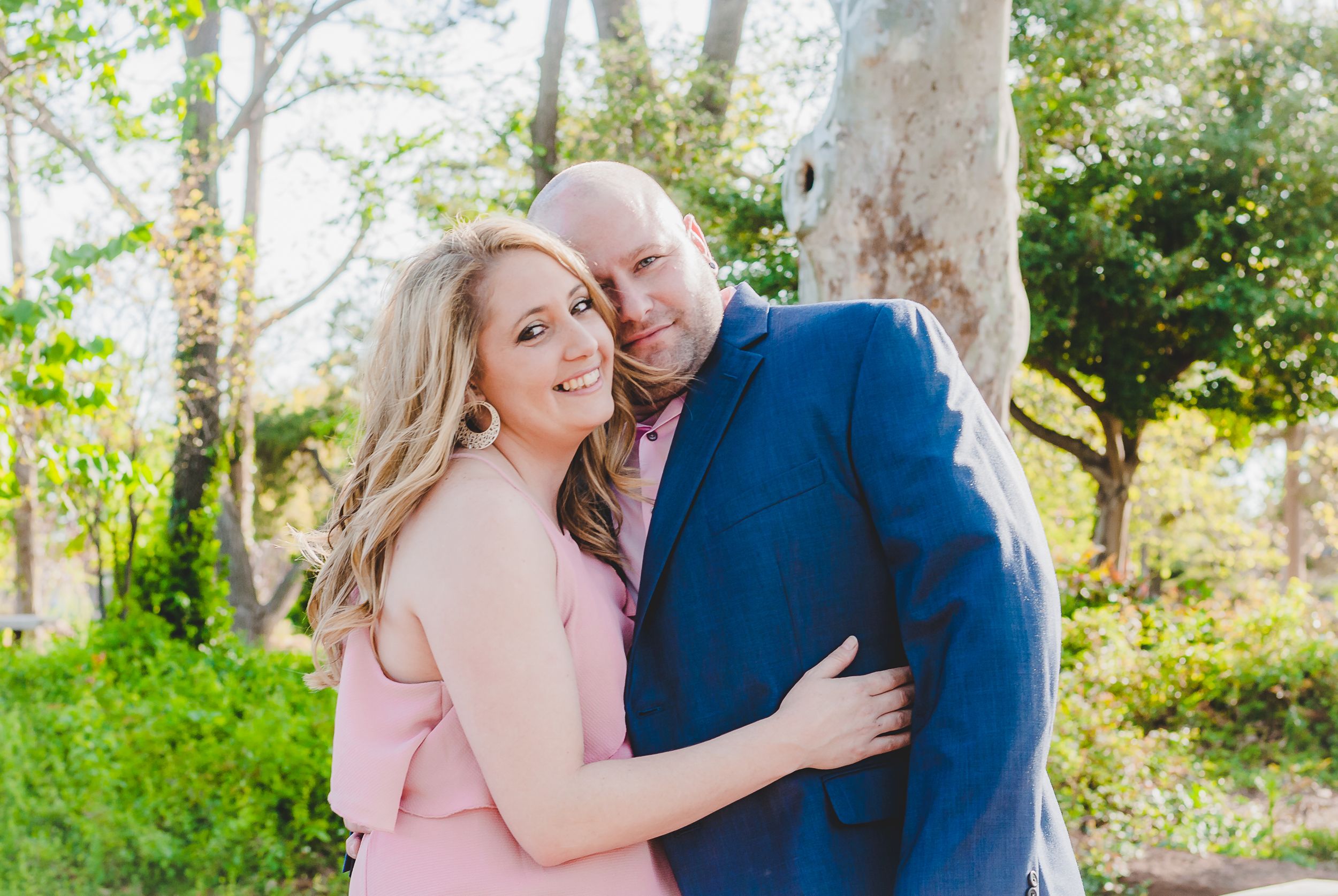 a portrait of a couple with the man and woman hugging each other in front of green scenery