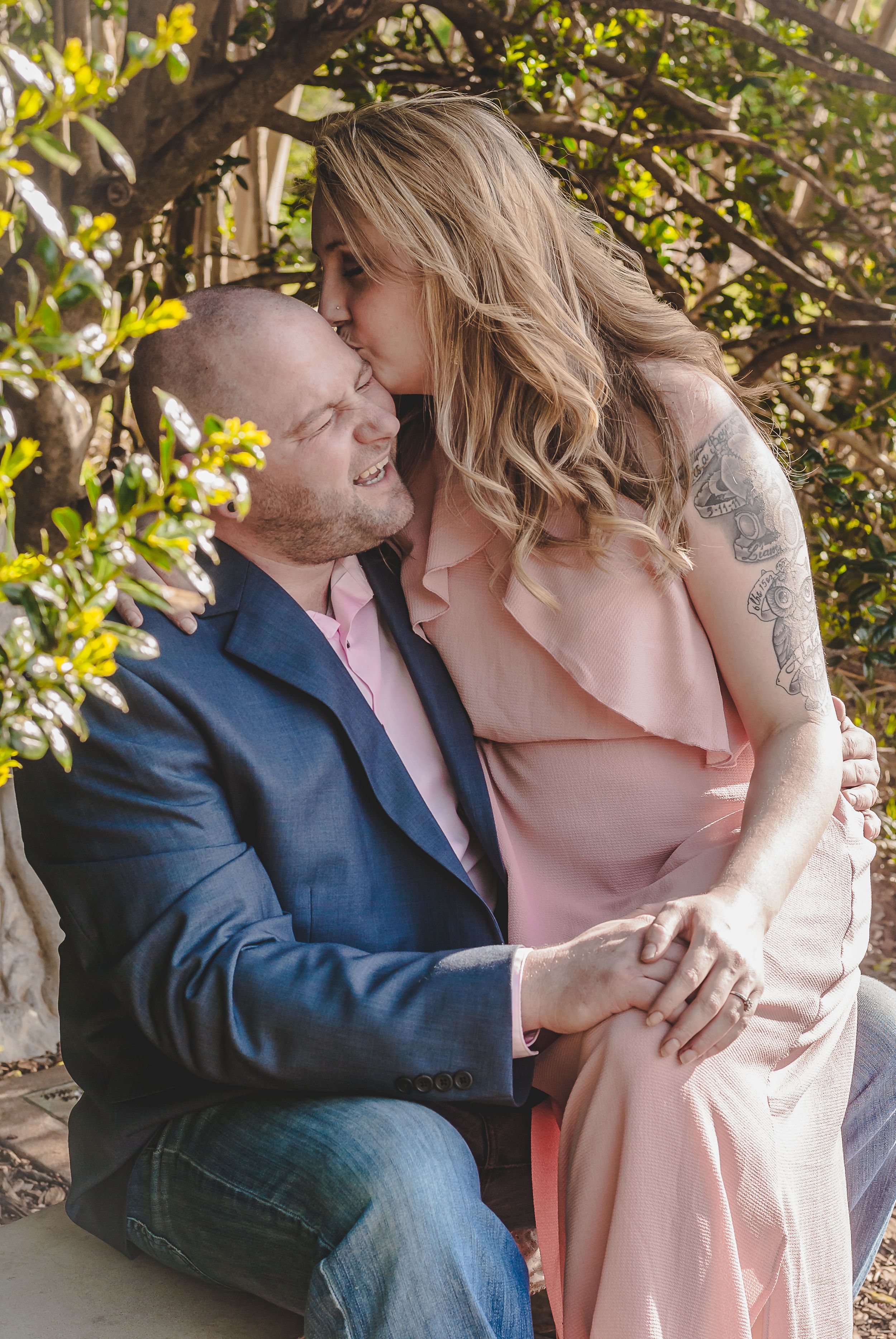 a portrait of a couple with the man sitting on a bench surrounded by greenery and the woman sitting on his lap kissing his forehead