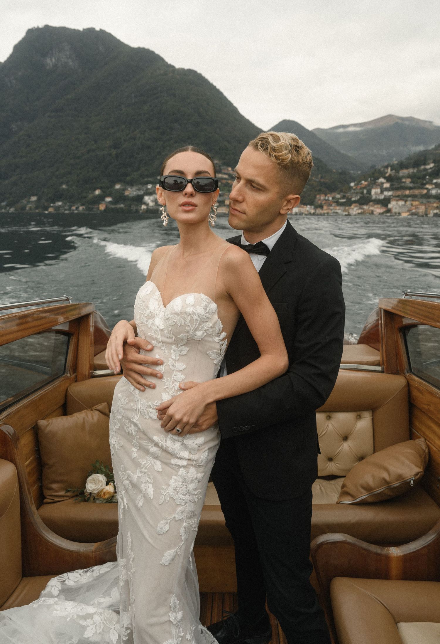 Stylish couple posing on luxury wooden boat with mountain lake views.